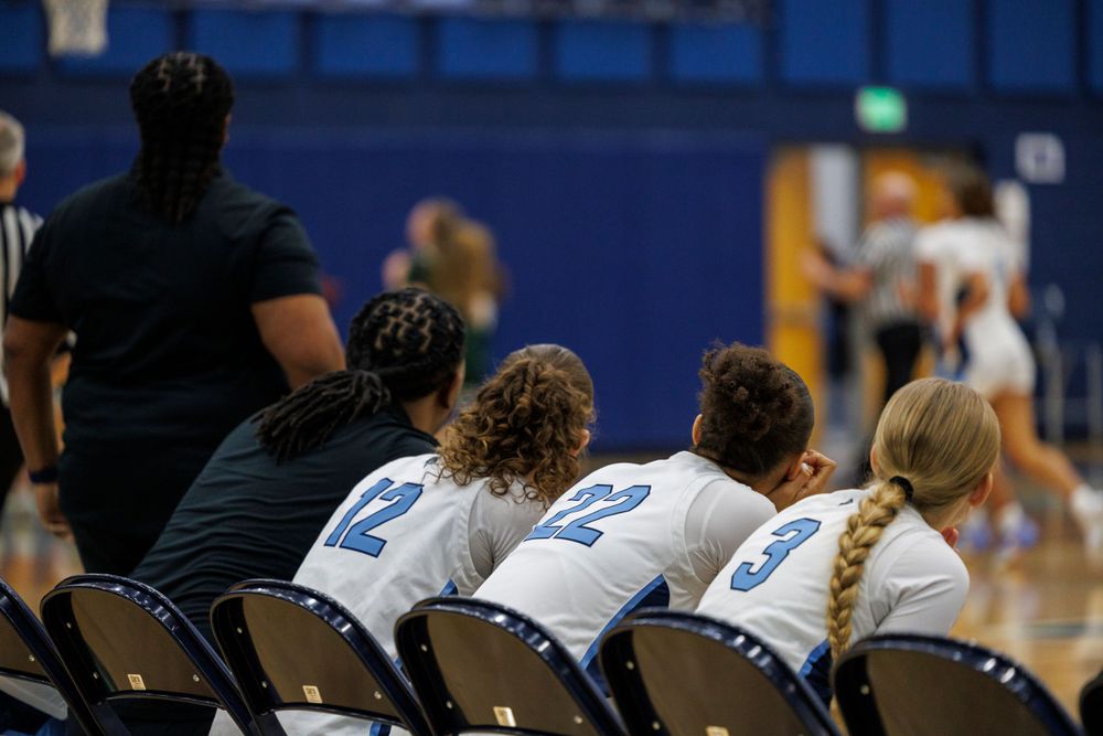 Players sit on sideline chairs watching basketball game in blue gymnasium.