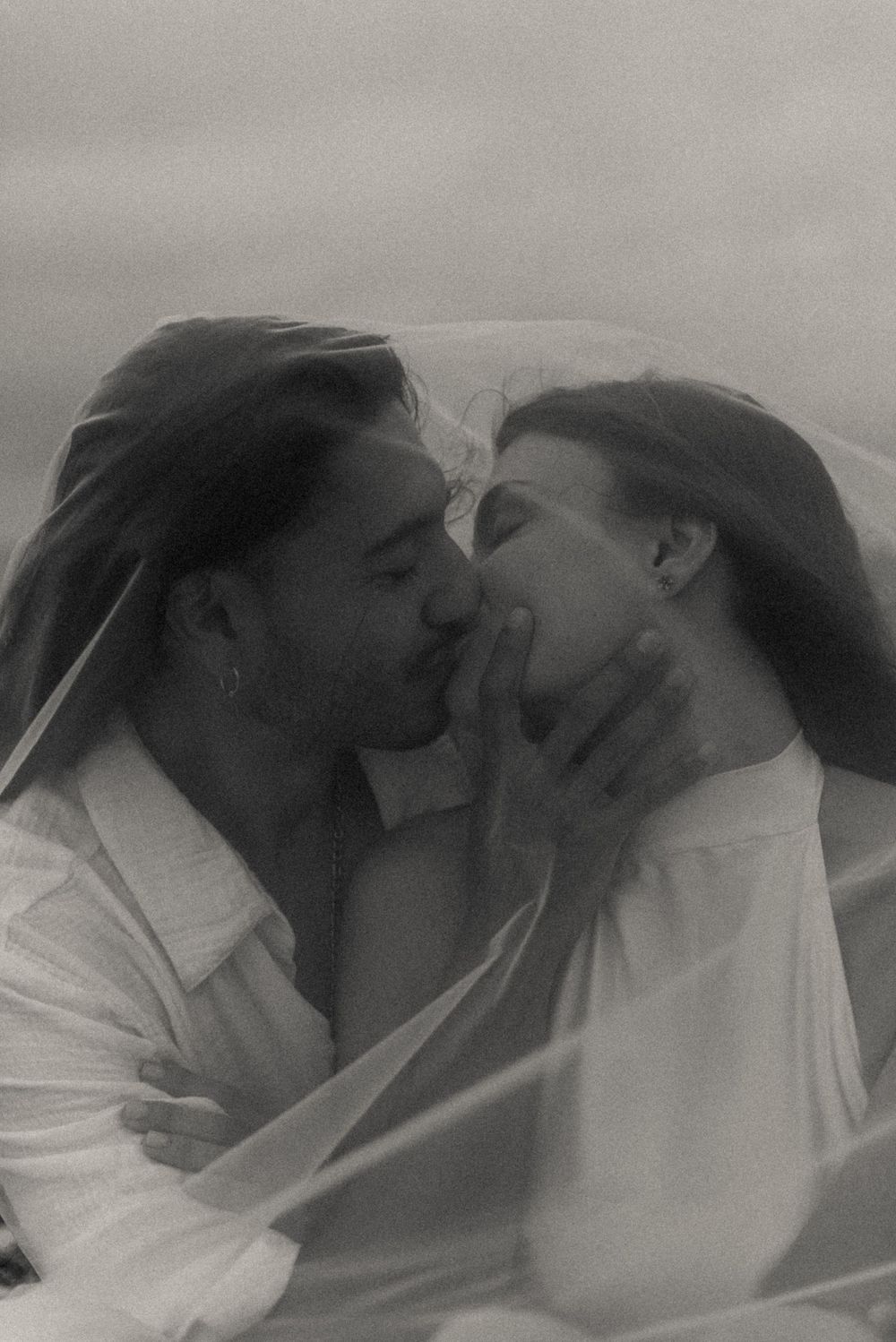 A black and white photo of a bride and groom kissing underneath her veil.