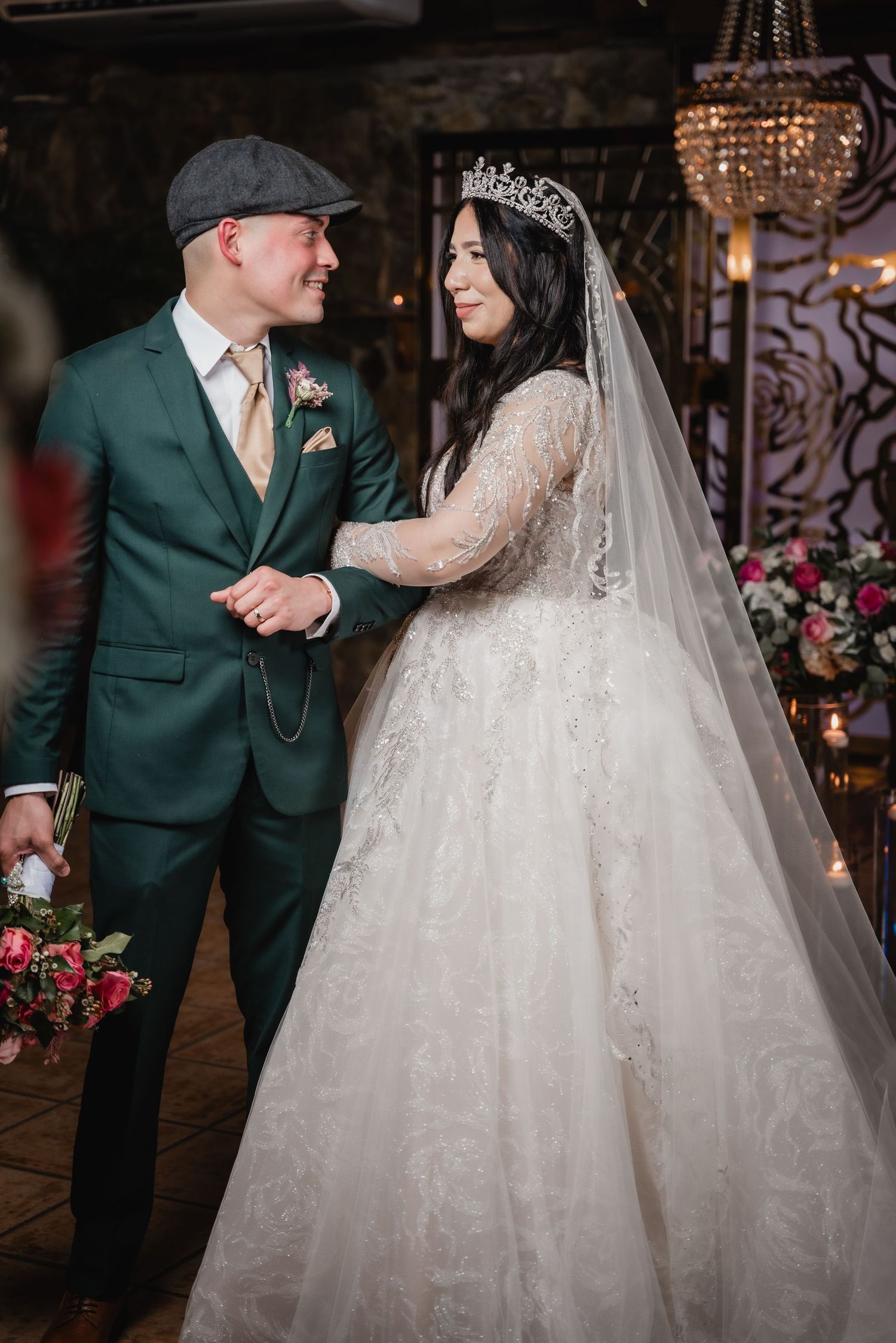 A newlywed couple in green suit and white lace gown share a romantic moment in an elegant venue with chandeliers.