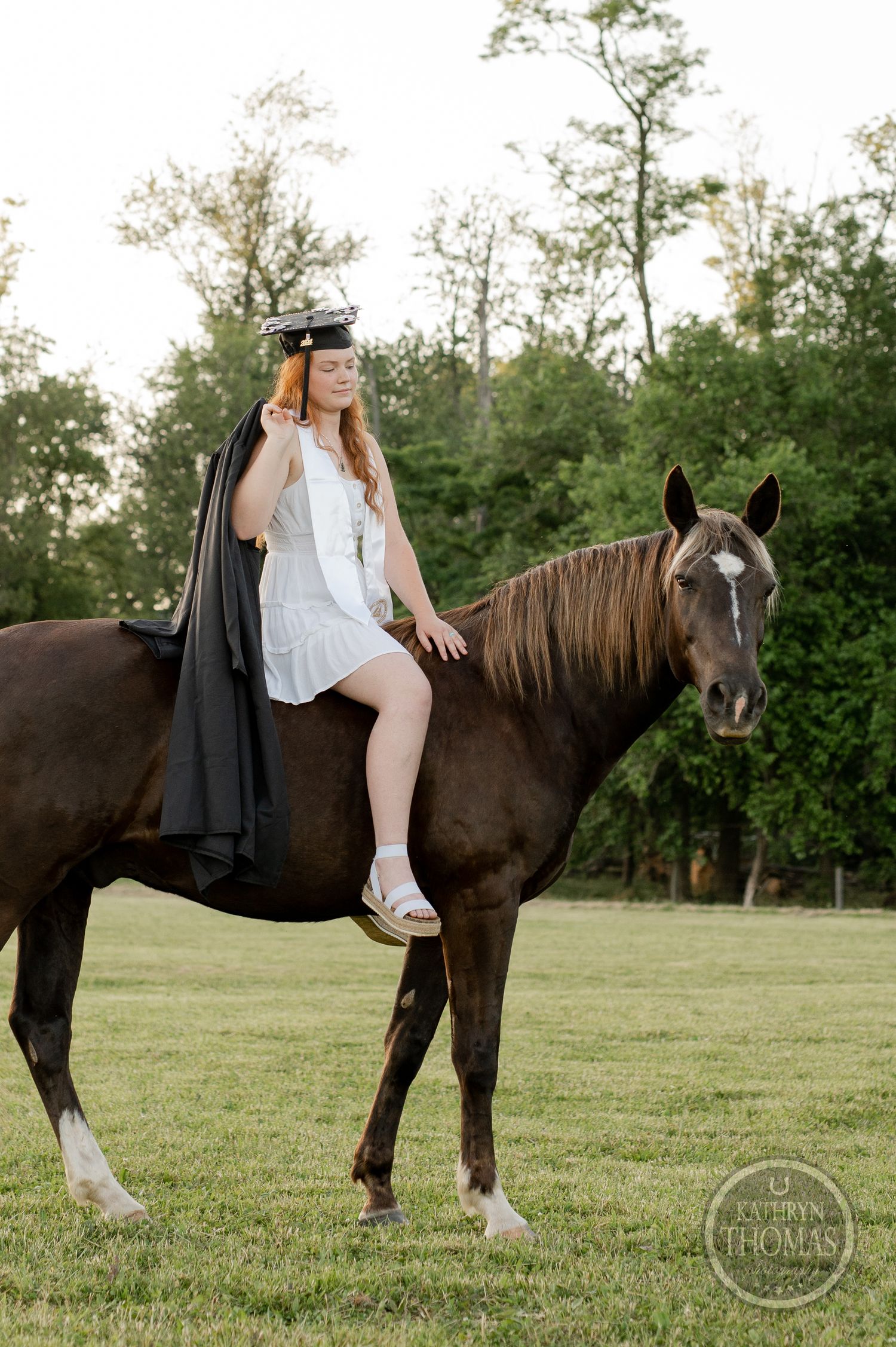 A graduate in academic regalia poses with a dark brown horse in a grassy field.