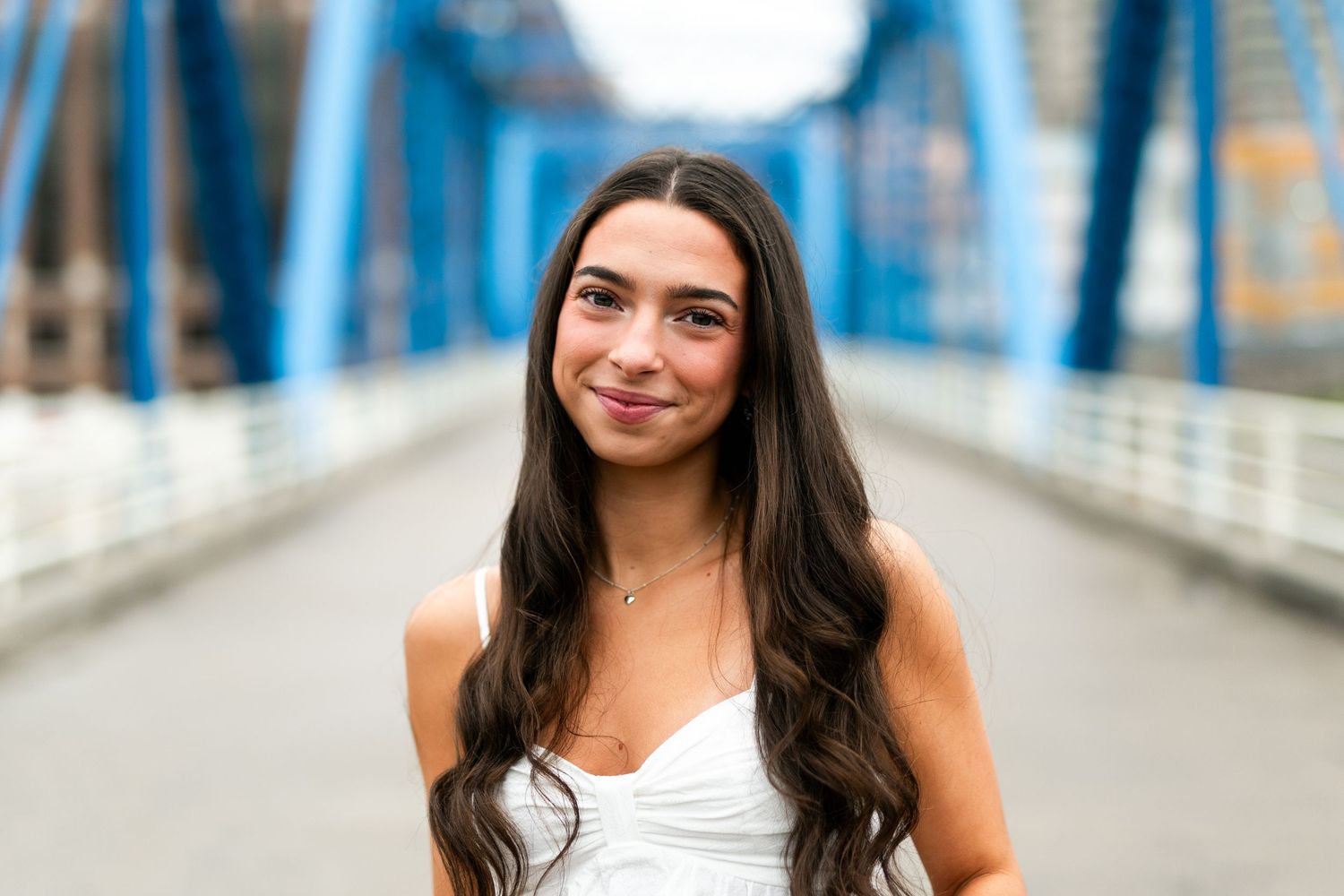 Someone with long dark hair and a white tank top smiles warmly while standing on a blue bridge walkway.