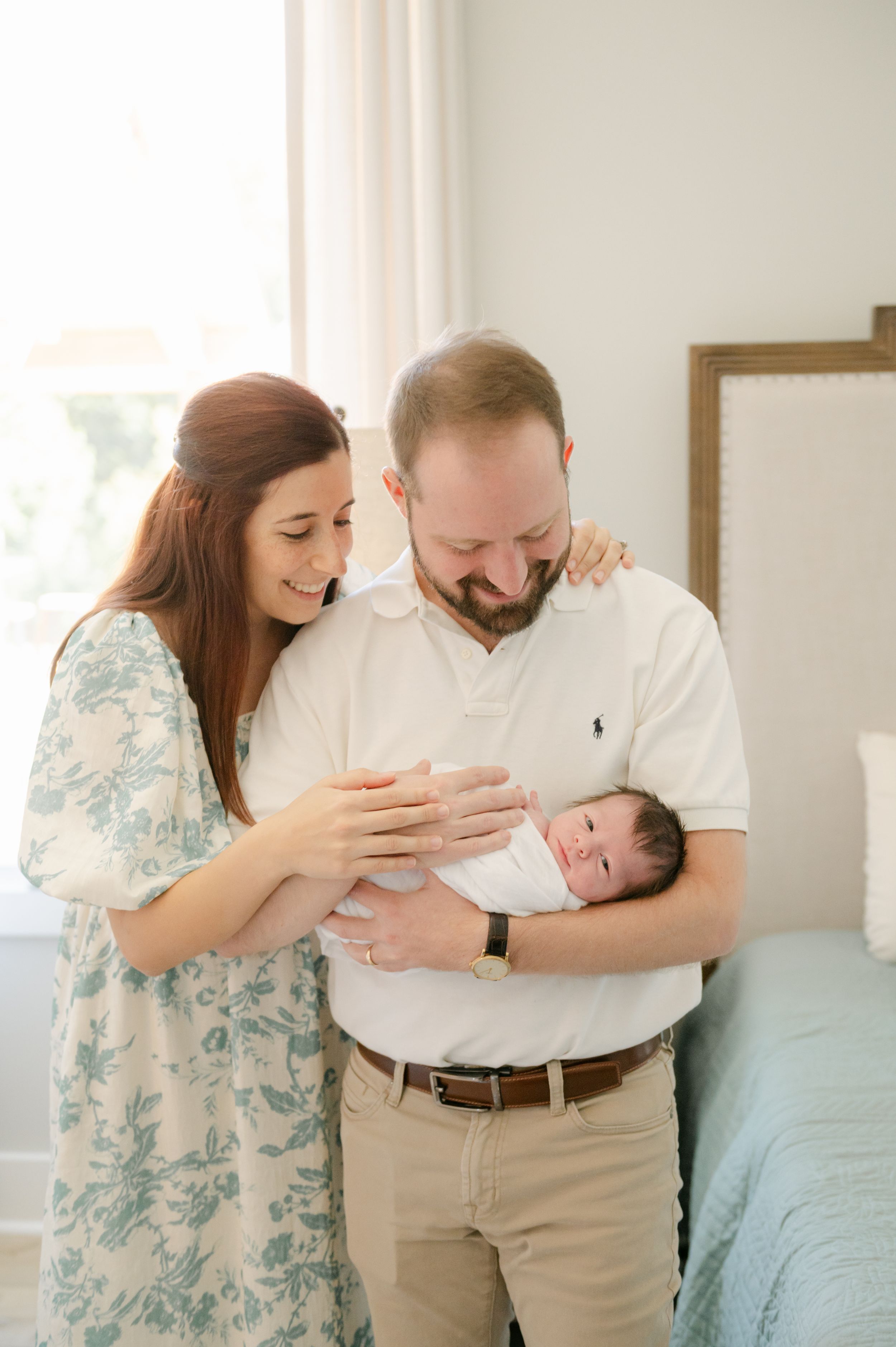 New parents smiling lovingly at swaddled newborn baby in bright bedroom setting.