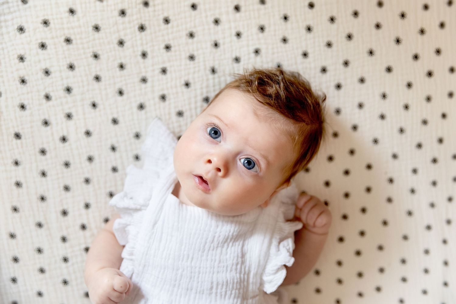Baby with big blue eyes laying on polka-dot crib sheet, looking up