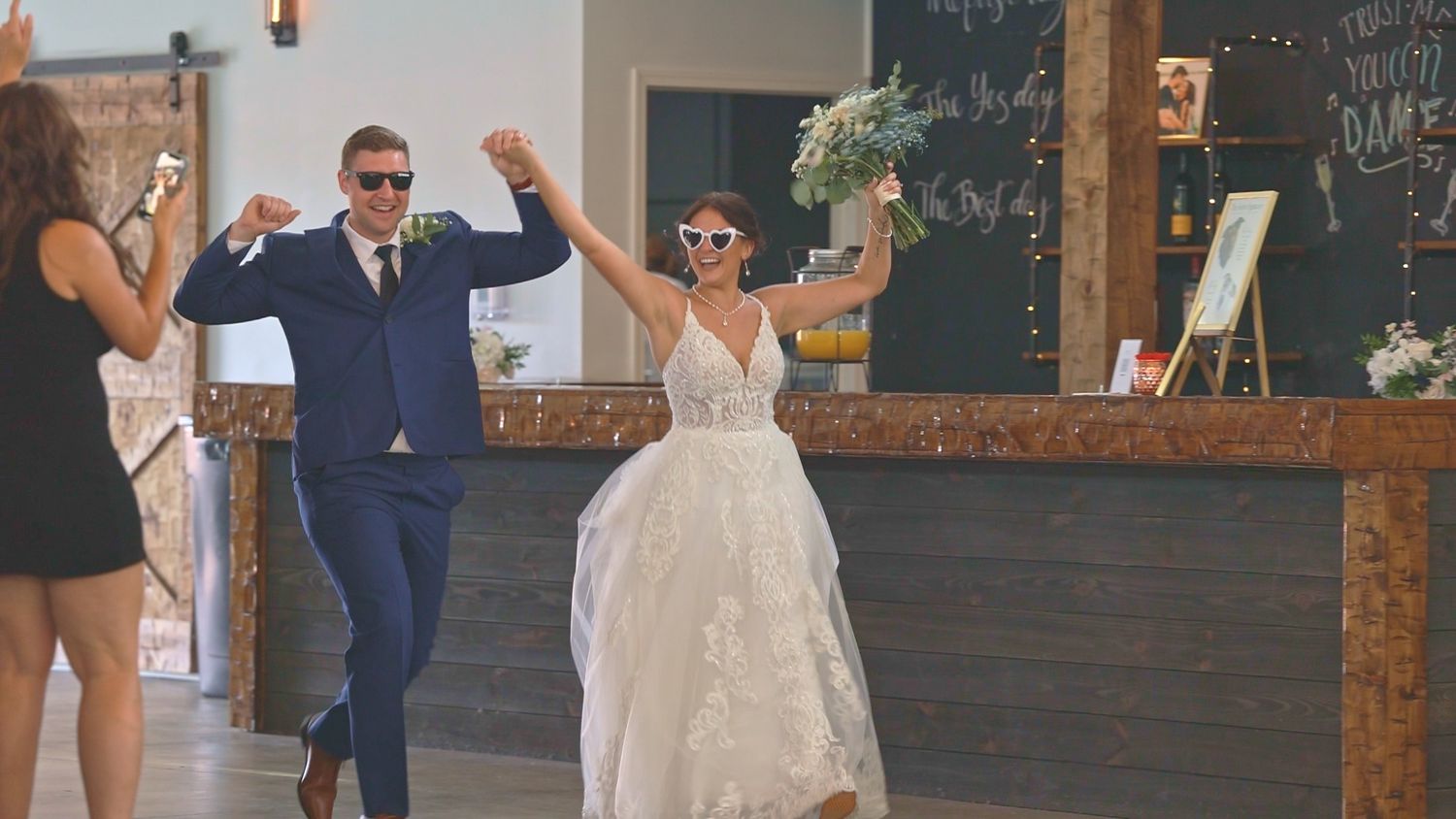 A bride and groom joyfully enter their wedding reception venue together with raised arms in celebration.