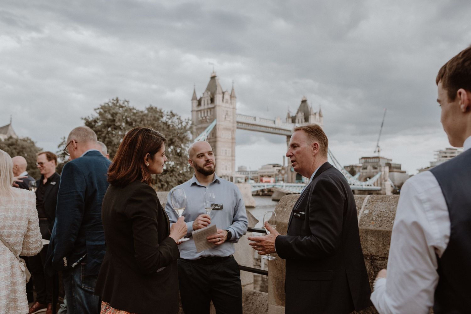 A group of people enjoying drinks at an outdoor gathering with London's iconic Tower Bridge visible in the background.