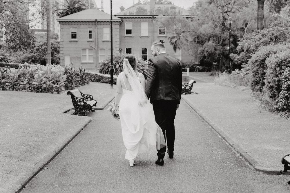 Black and white photo of a couple in wedding attire walking away, captured by Apertured Media Auckland Wedding Photographer.