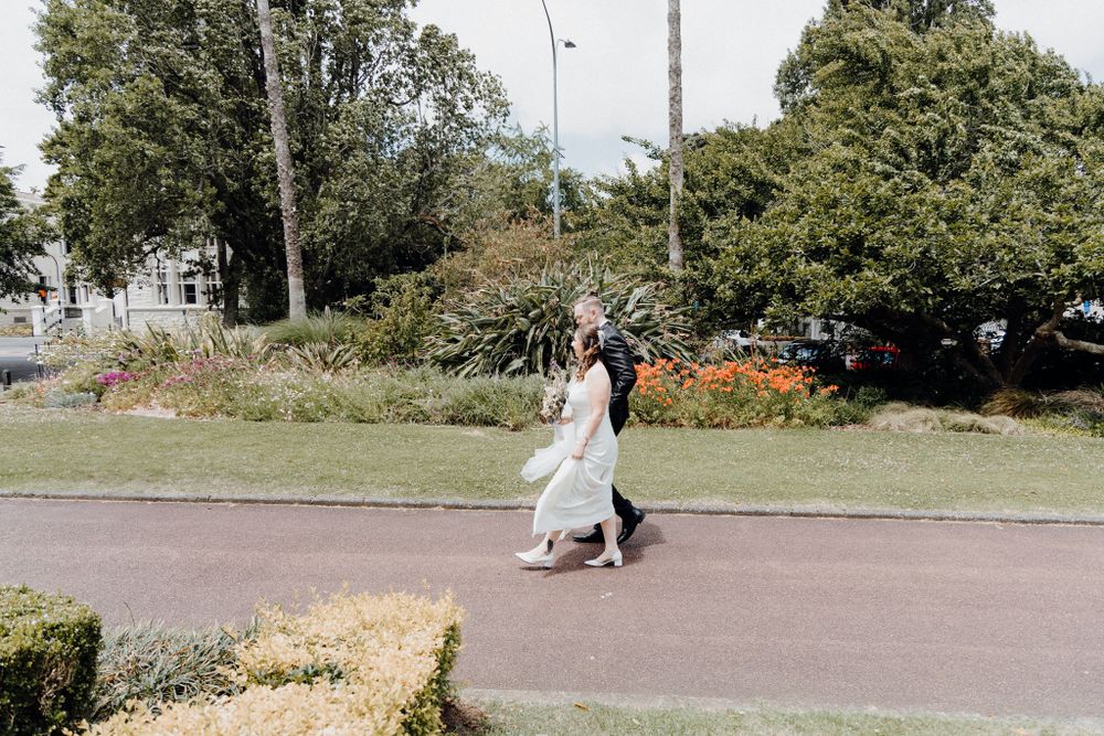 A couple walks together on a paved path surrounded by gardens and greenery, captured by Apertured Media Auckland Wedding Photographer.