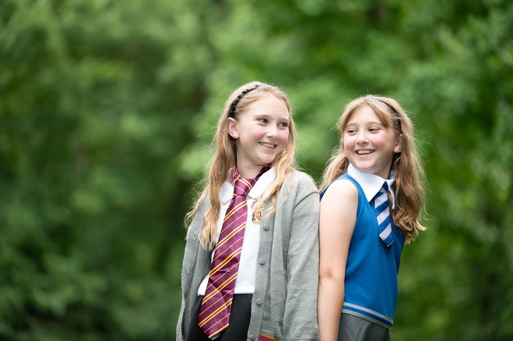 Two students in school uniforms with ties smile outdoors in a natural setting.
