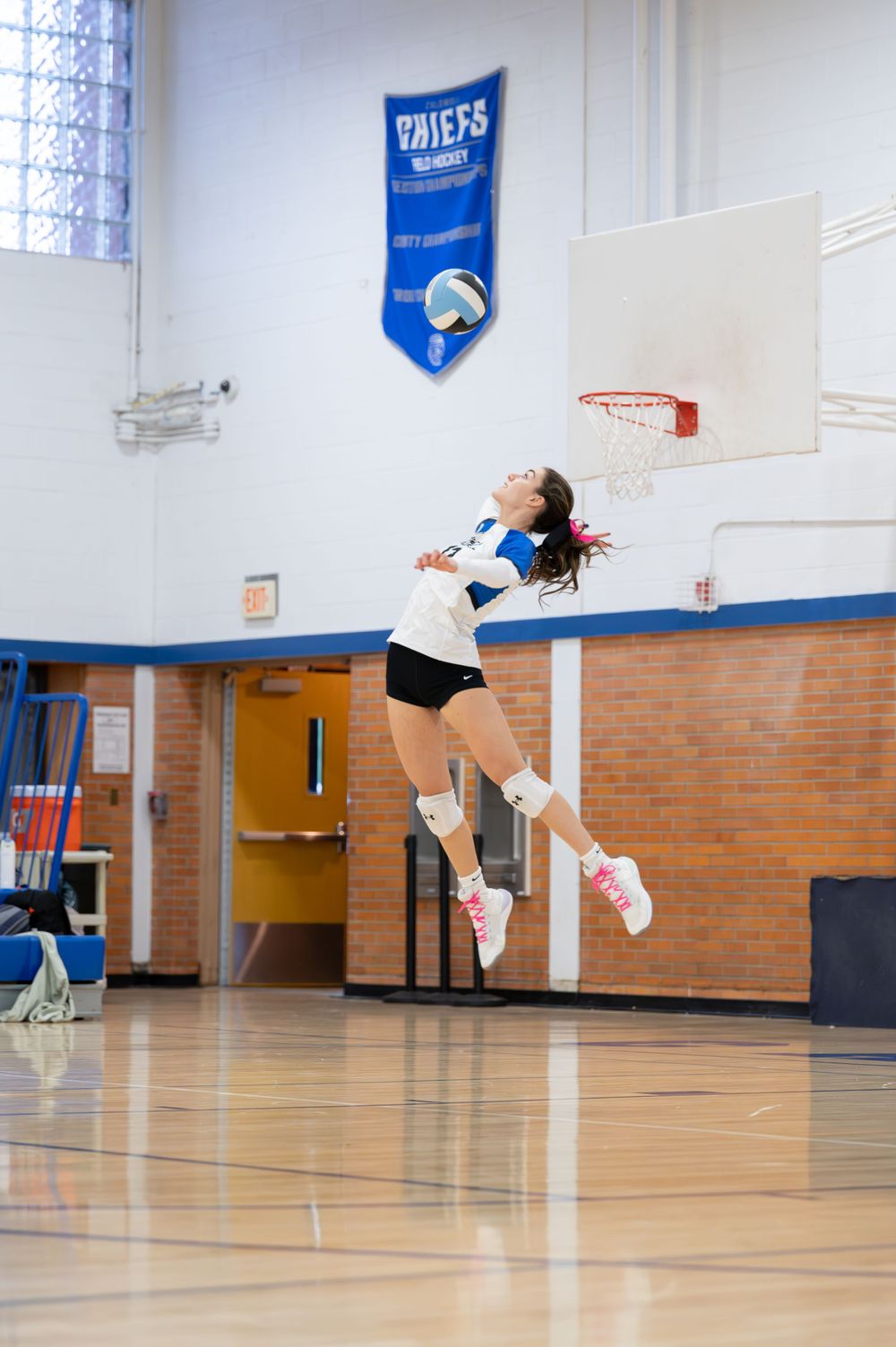 Female volleyball player jumping high to serve ball in school gymnasium.