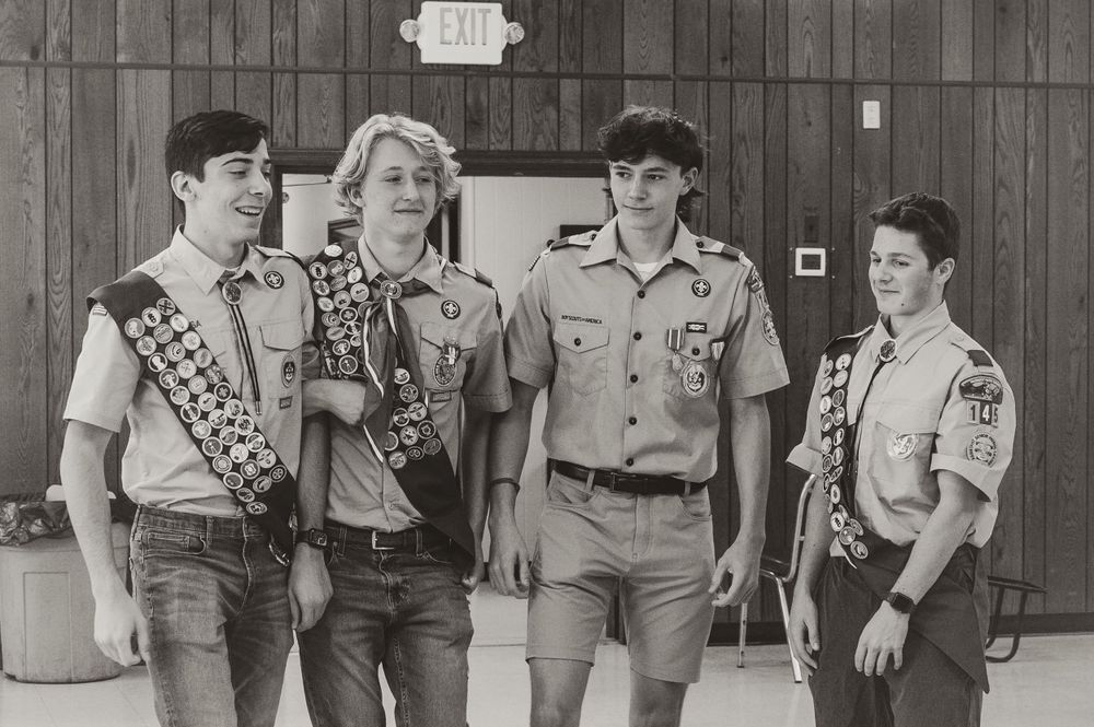 A group of Eagle Scouts wearing merit badge sashes pose together against a wooden wall background.