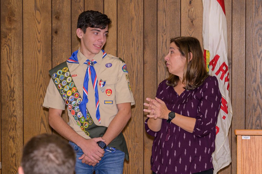 A Scout in uniform with merit badges speaks with an adult during a ceremony.