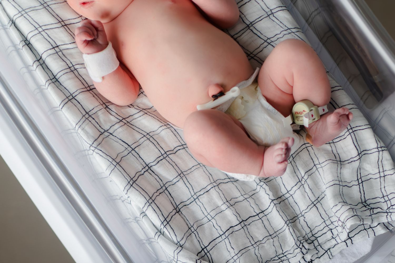 Baby wearing white diaper lies on a gray and white plaid changing pad with soft lighting from above.