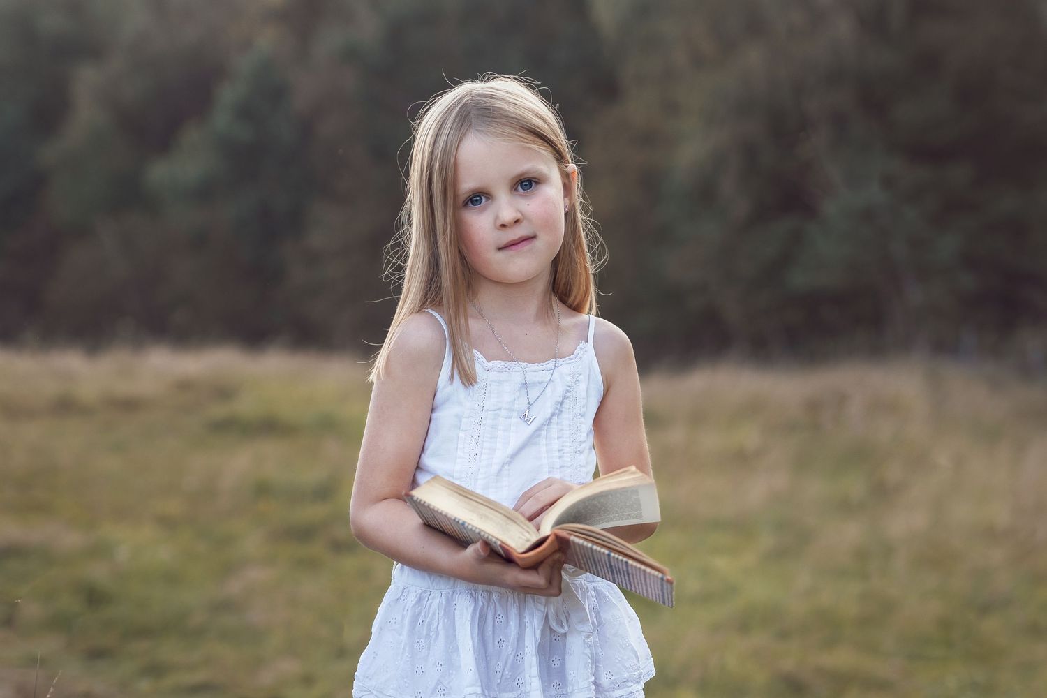 En flicka står vid en äng, med skog i bakgrunden. Flickan håller en bok i sina händer.