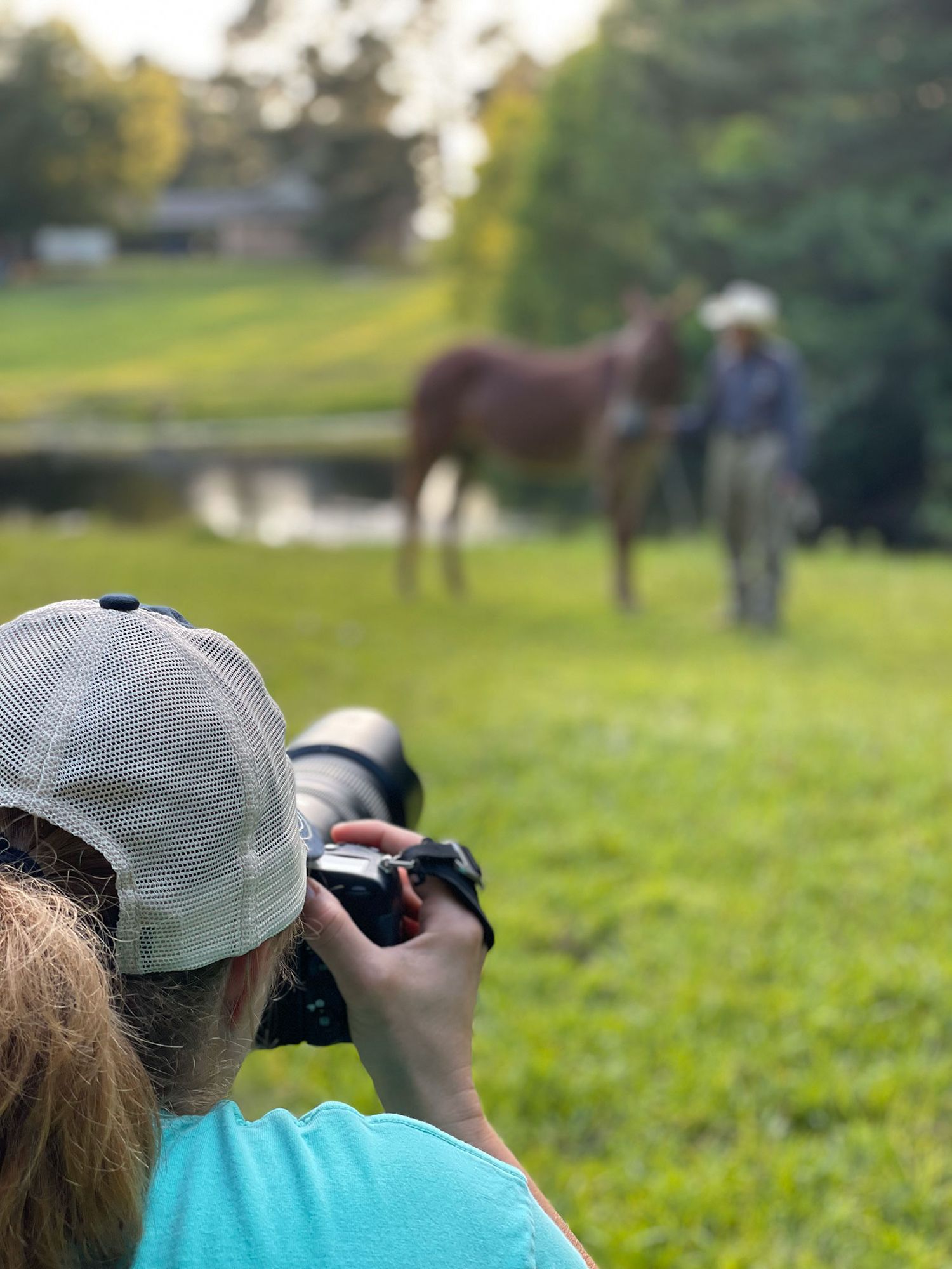 Photographer wearing turquoise shirt and cap captures image of horse and handler by pond in pastoral setting.