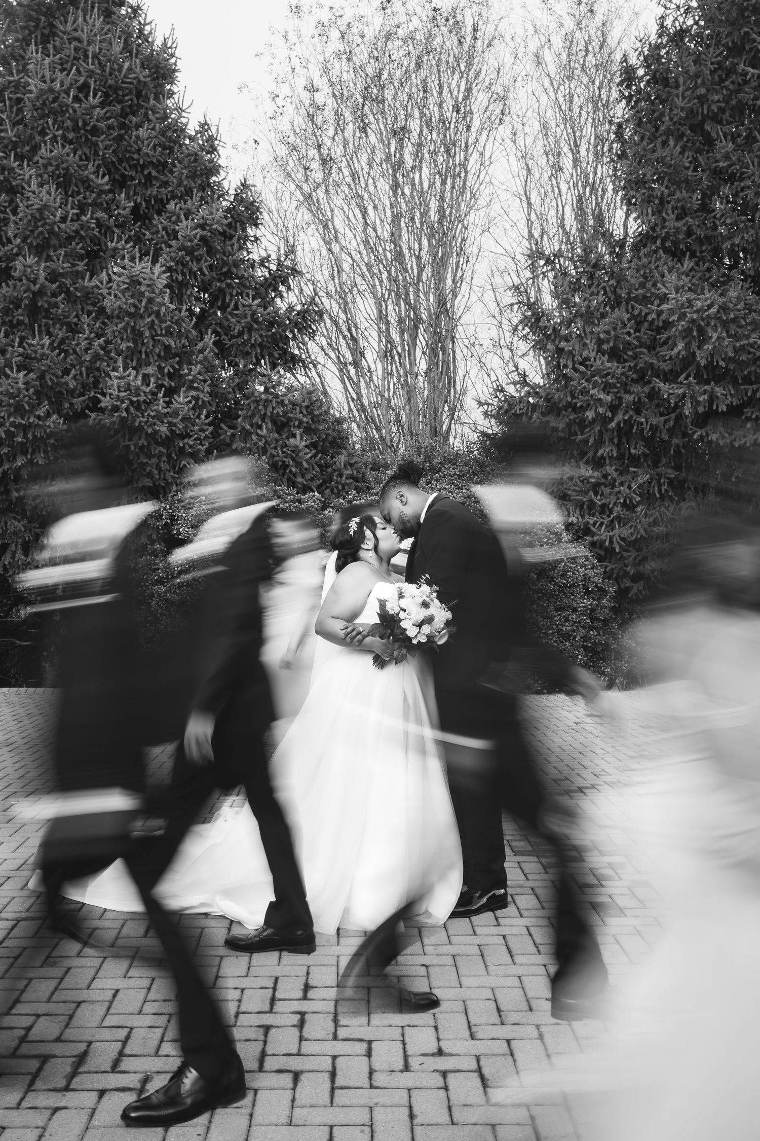 Black and white motion blur photo of bride and groom kissing as wedding party walks past.