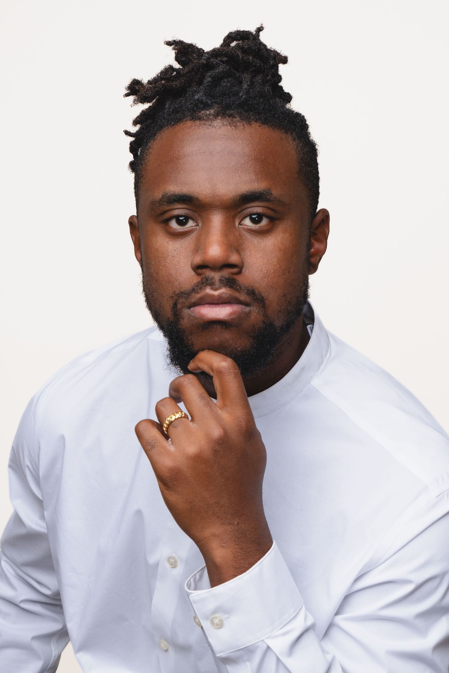 A confident Black man with dreadlocks and a beard poses thoughtfully in a white shirt, resting his chin on his hand.