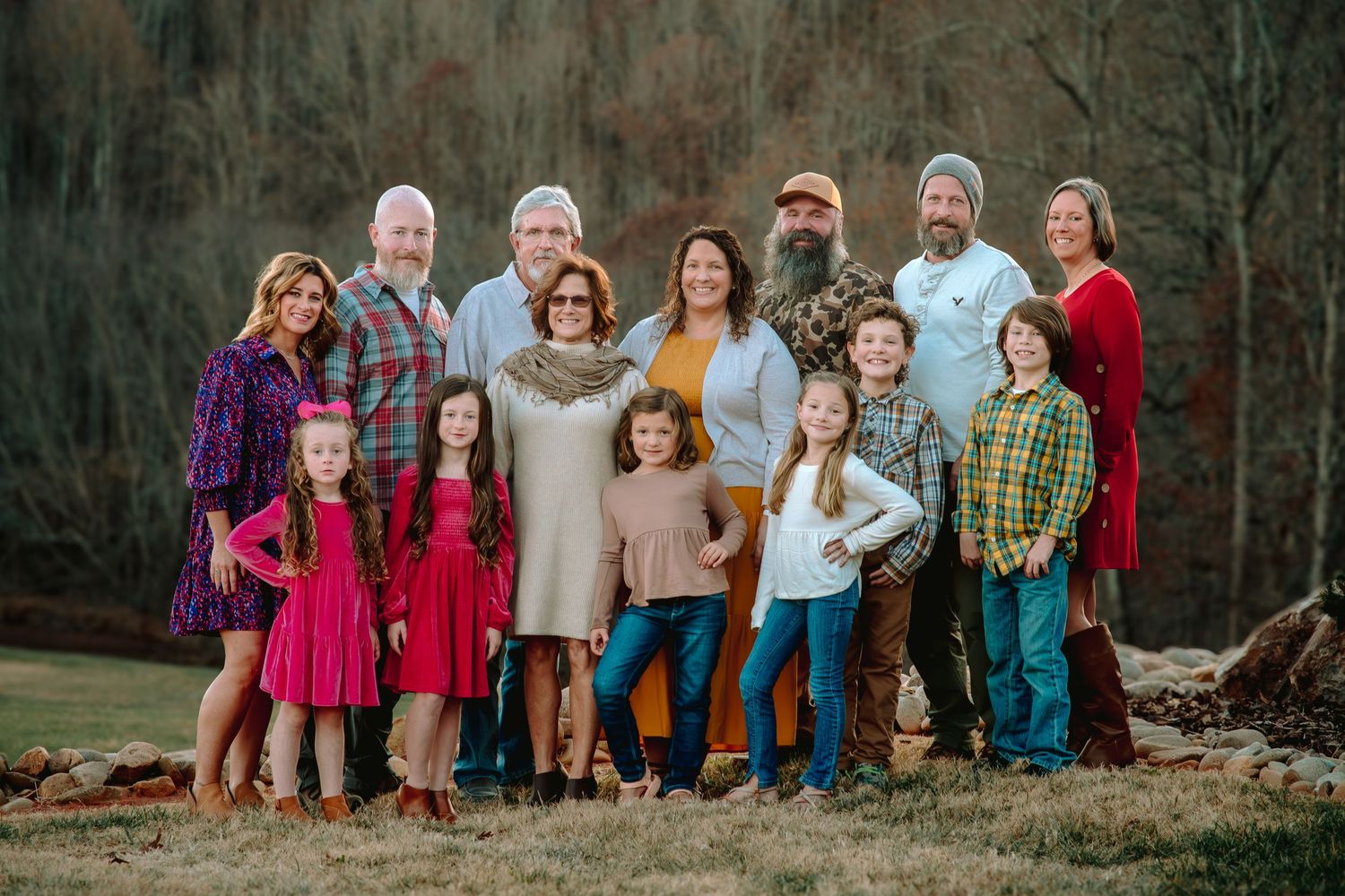 Large multi-generational family poses together outdoors against wooded backdrop during fall season photo session.