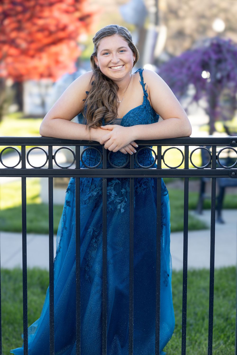Someone wearing an elegant navy blue lace dress poses against an iron railing with colorful autumn trees in the background.