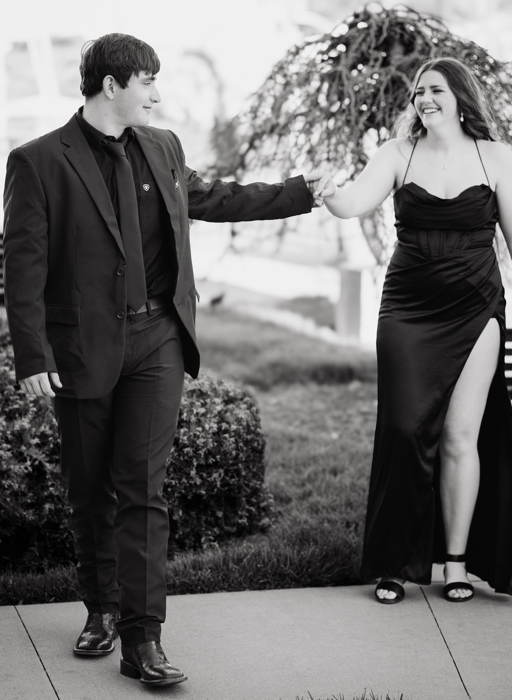 A couple in formal attire share a playful moment outdoors in black and white photography.