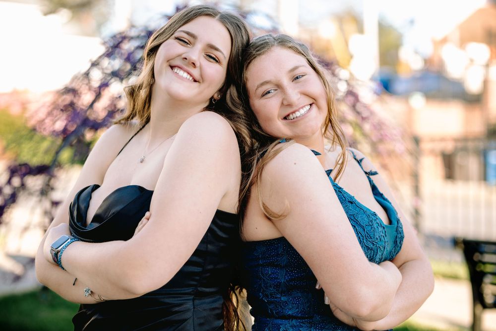 Two friends in formal dresses stand back-to-back smiling outdoors during golden hour.
