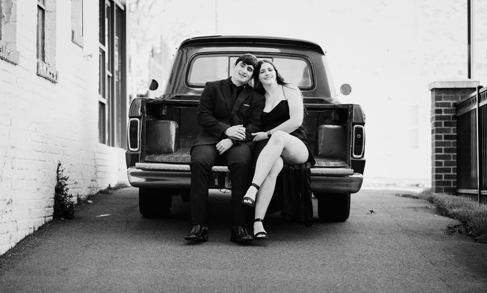 A couple poses on the tailgate of a vintage pickup truck in black and white photography.