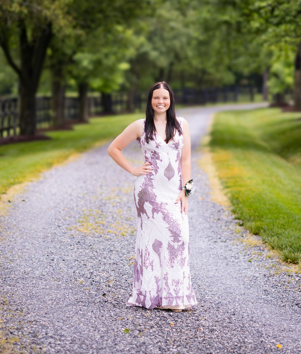 Woman in floral maxi dress poses on gravel path lined with trees and grass.