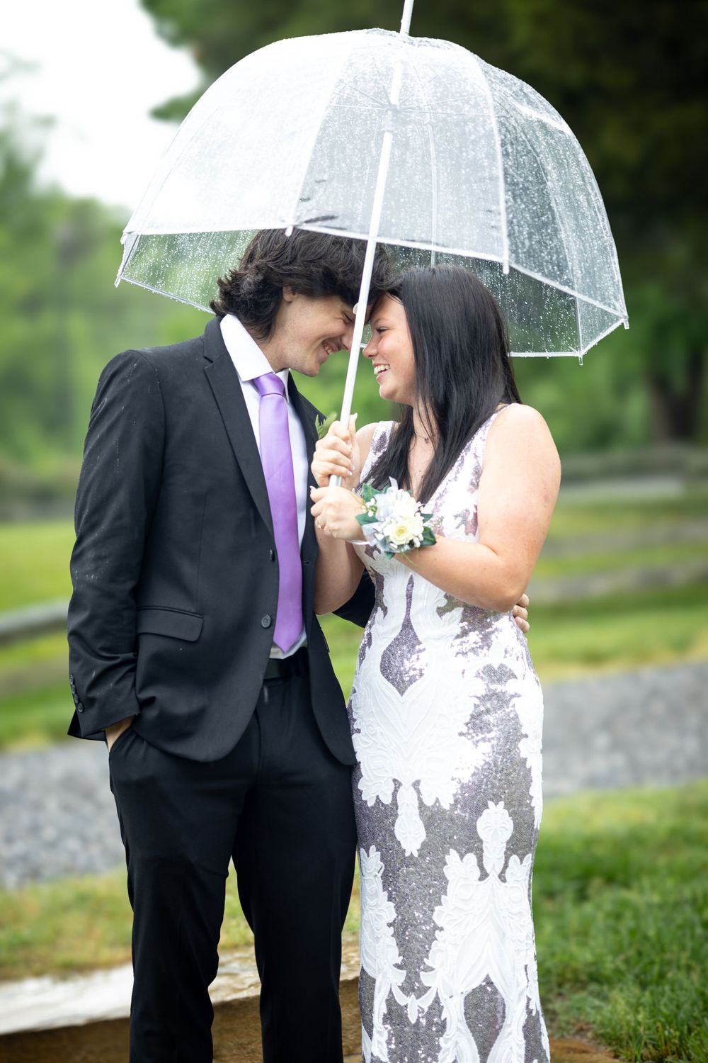 Couple sharing clear umbrella in formal attire during light rain.
