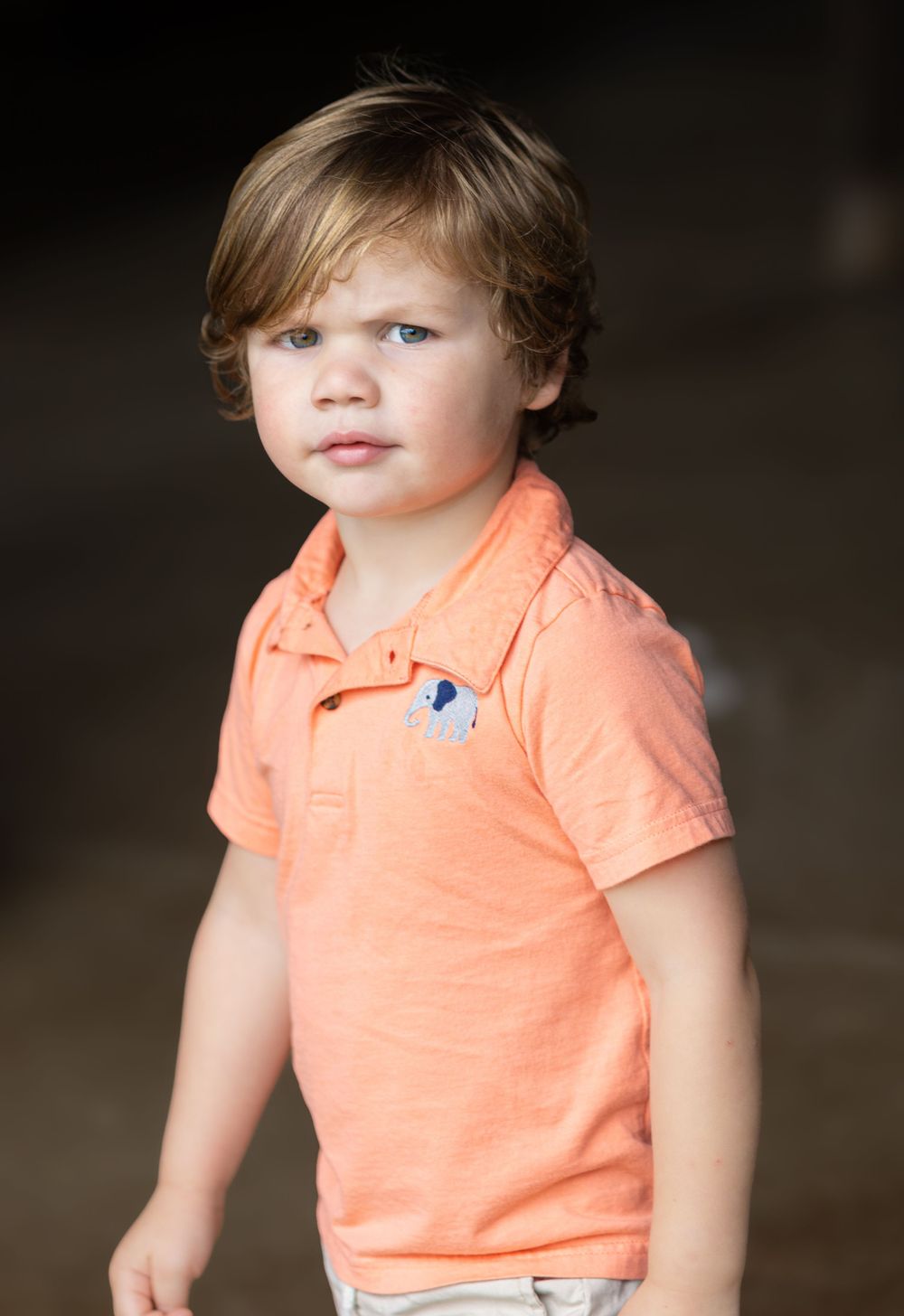 Young child wearing coral colored polo shirt against dark background.