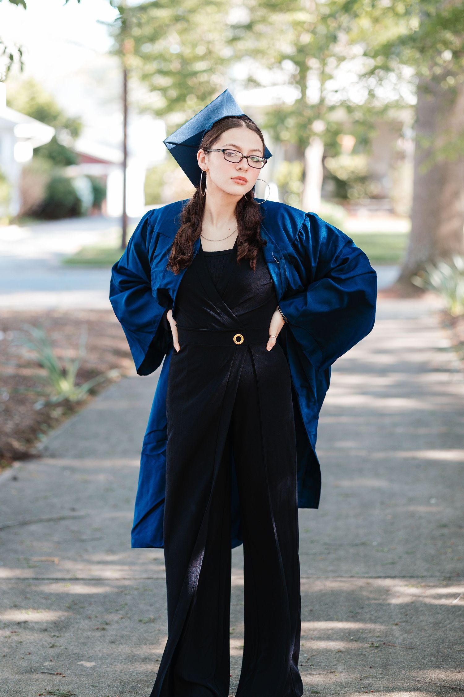 Graduate in a blue academic gown and black dress posing on a sidewalk.
