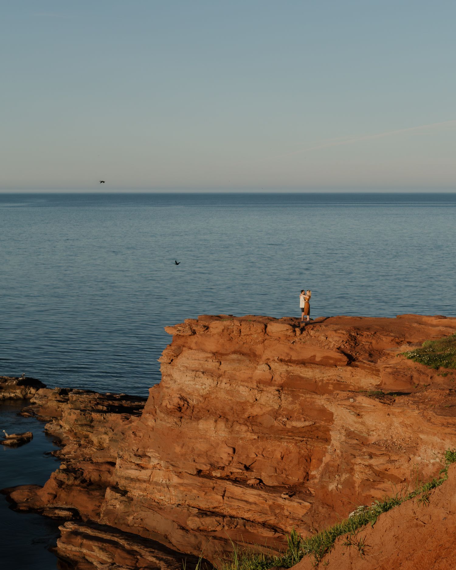 Figure stands on dramatic red cliff overlooking vast ocean during golden hour.