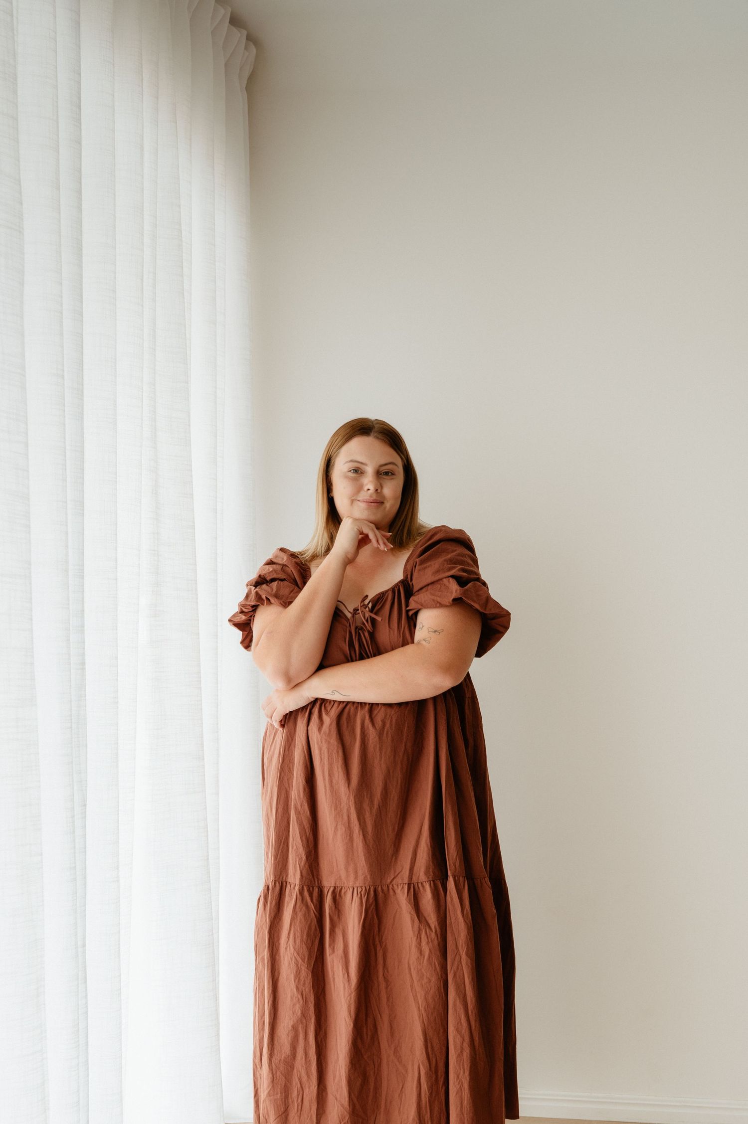 Person wearing black sleeveless top and white pants sits casually against white wall with sandals.