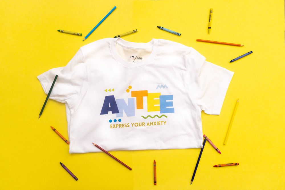 White t-shirt with colorful text design displayed on vibrant yellow background with scattered colored pencils around it.