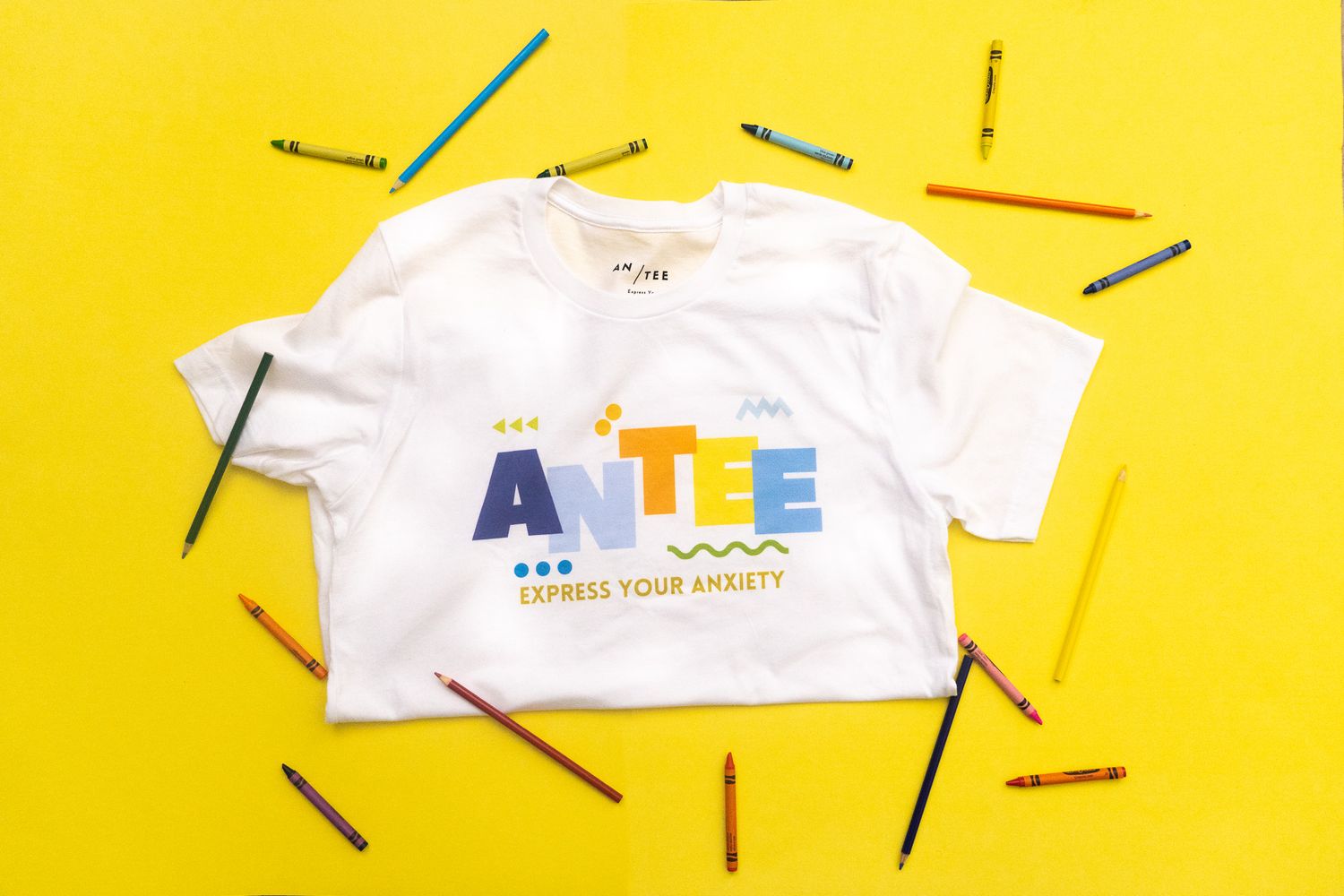 White t-shirt with colorful text design displayed on vibrant yellow background with scattered colored pencils around it.
