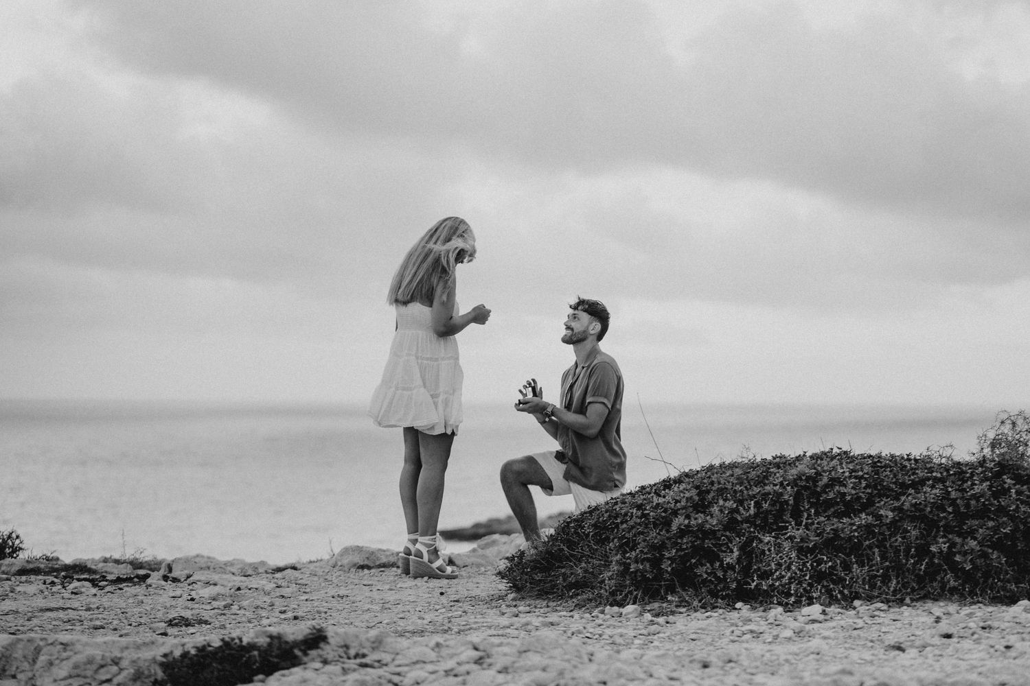 A romantic marriage proposal scene on a rocky coastal overlook in black and white photography.