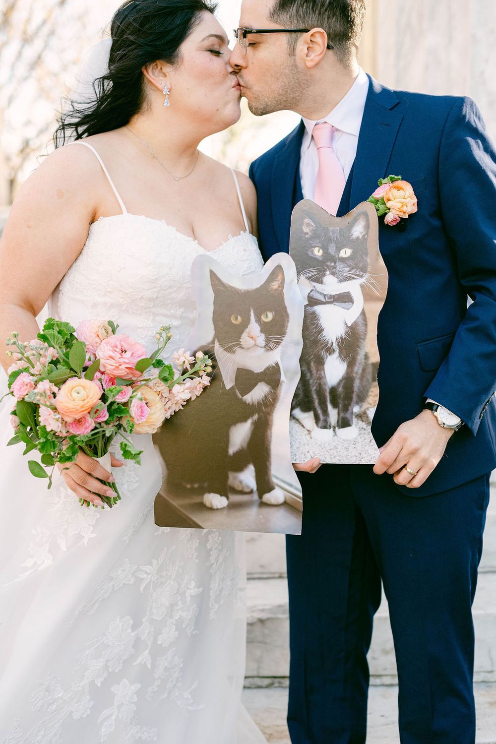Wedding couple embracing while holding a pink floral bouquet against a light background.