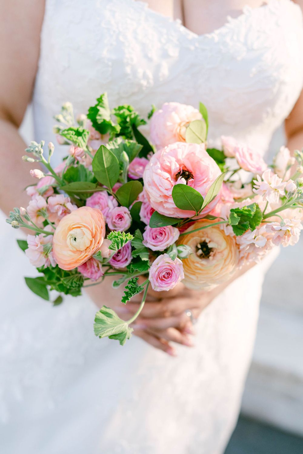 A delicate bridal bouquet featuring pink ranunculus, roses, and greenery held against a white wedding dress.