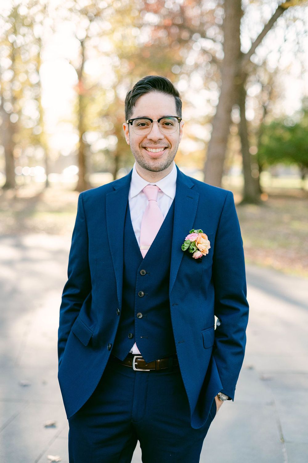 A groom in a navy blue three-piece suit and pink tie stands outdoors on an autumn day.