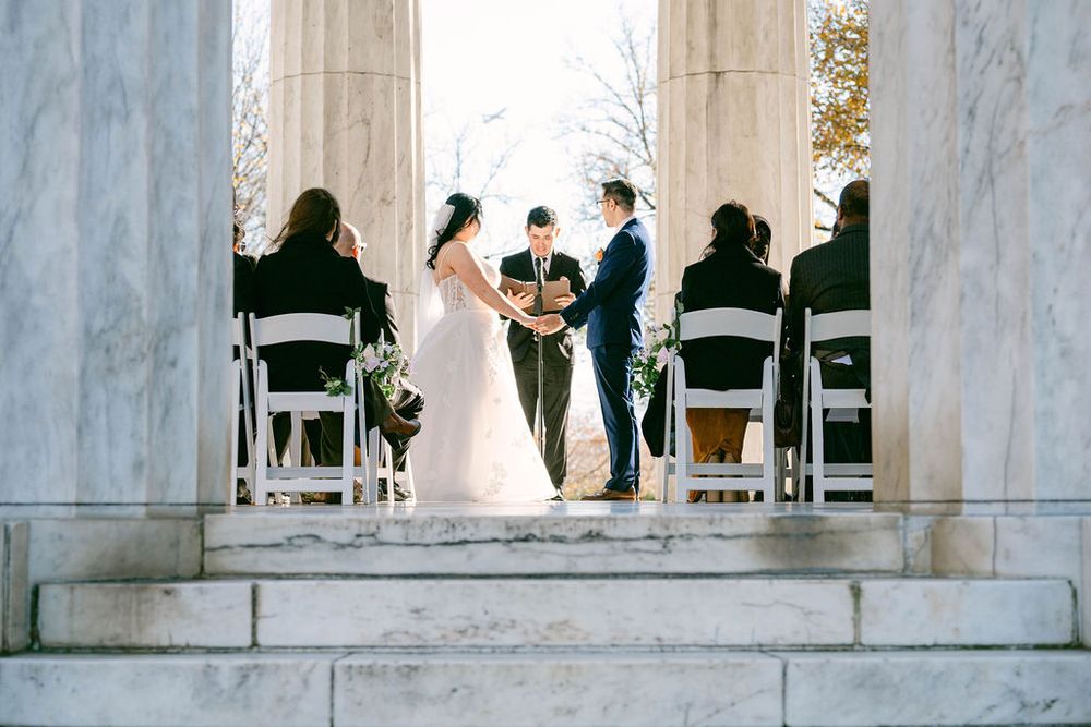 Wedding ceremony takes place in an elegant white marble venue with guests seated in black chairs.