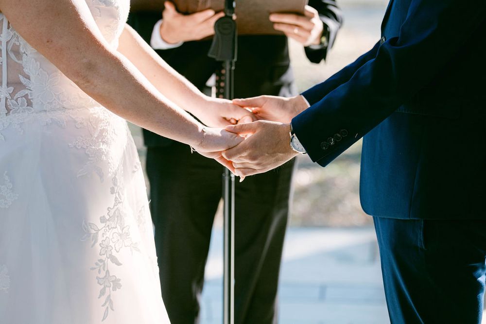 Close-up shot of hands clasped together during an outdoor wedding ceremony with an officiant in the background.