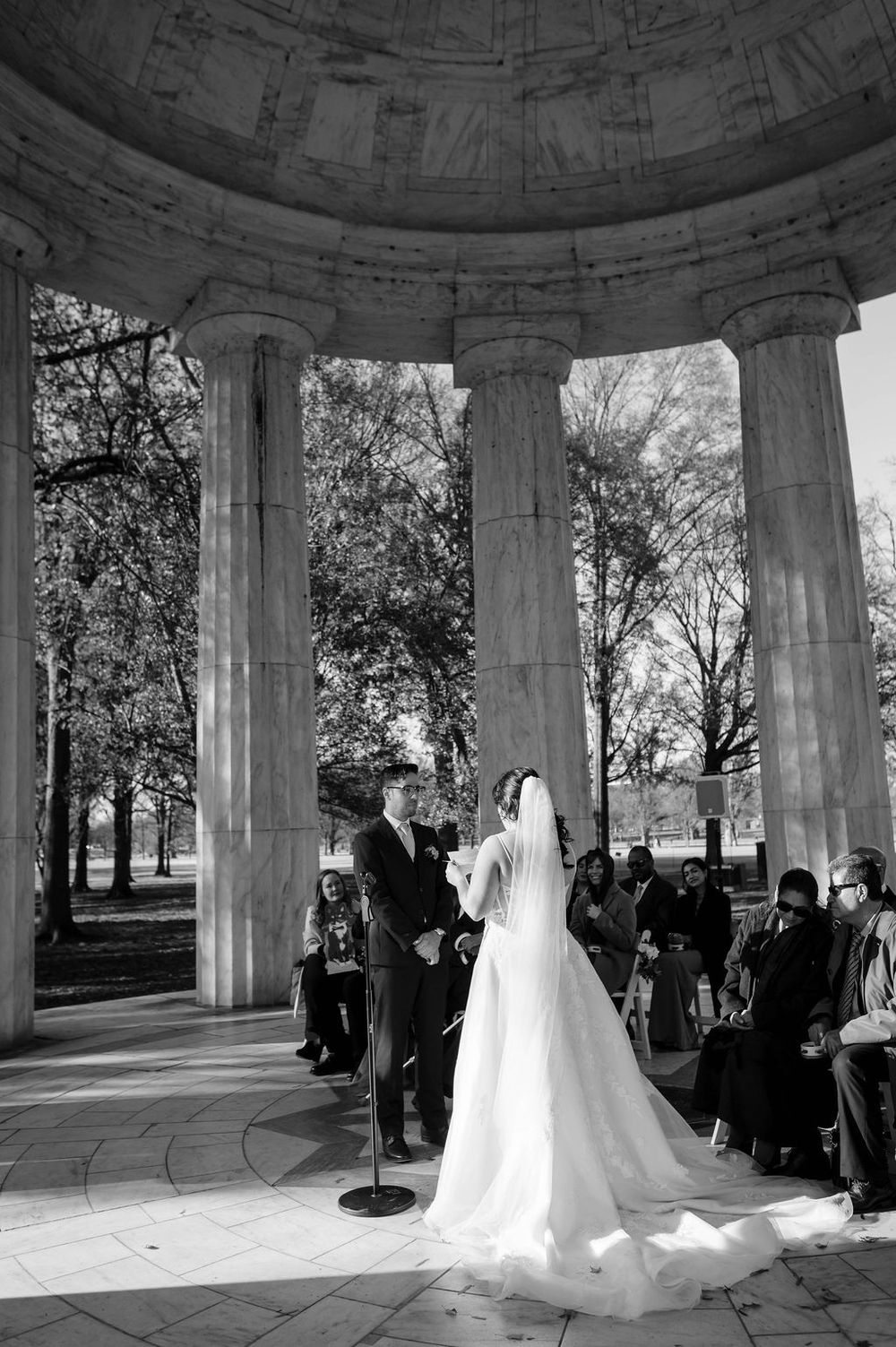 A wedding ceremony takes place in a classical stone gazebo surrounded by autumn trees in a park setting.