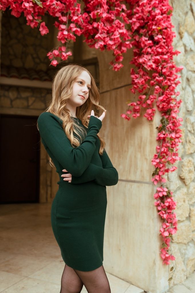 A person in an emerald sweater dress poses beneath an archway covered in bright pink flowering vines.