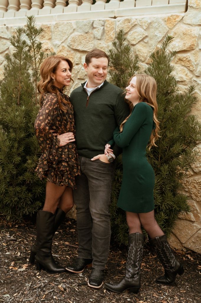 Three people posing together in front of evergreen trees and a stone wall.