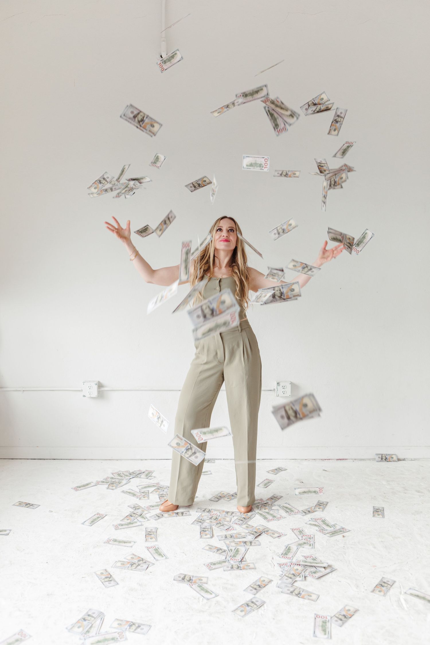 Someone in beige overalls celebrates joyfully while money bills float and fall around them in a white studio setting.