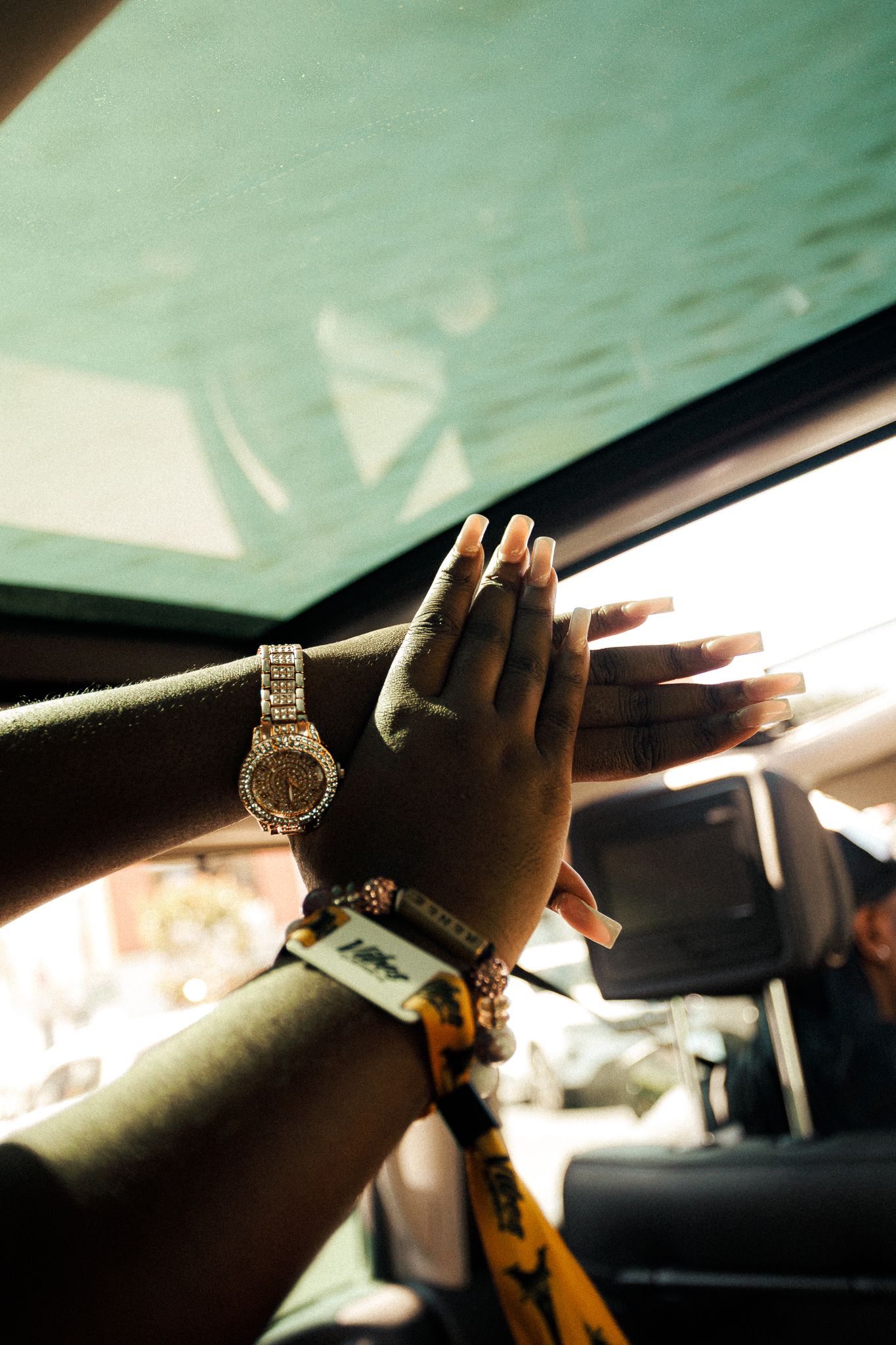 A hand wearing a gold watch and jewelry is silhouetted against a car window in warm sunlight.