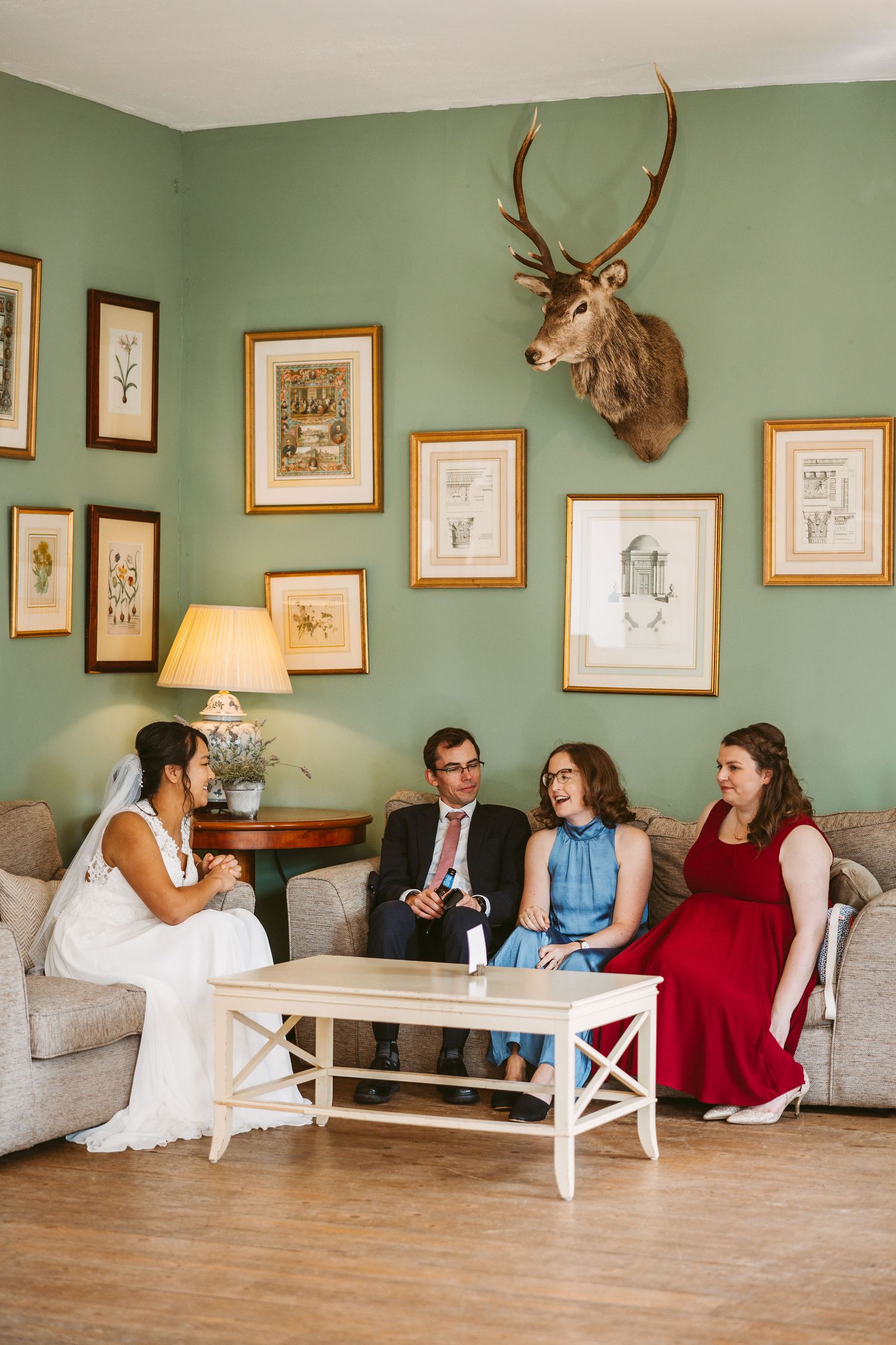 Wedding guests relax in an elegant sitting room with sage green walls adorned with framed artwork and mounted deer antlers.