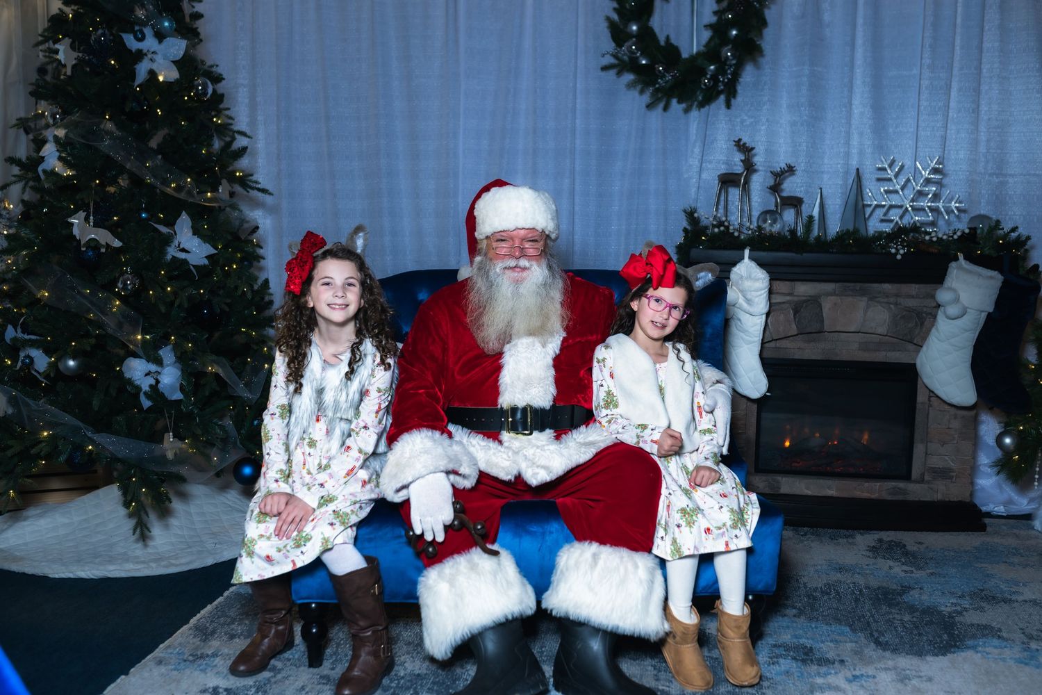 Traditional Christmas scene with Santa in red suit seated by decorated tree and fireplace with two children in matching dresses.