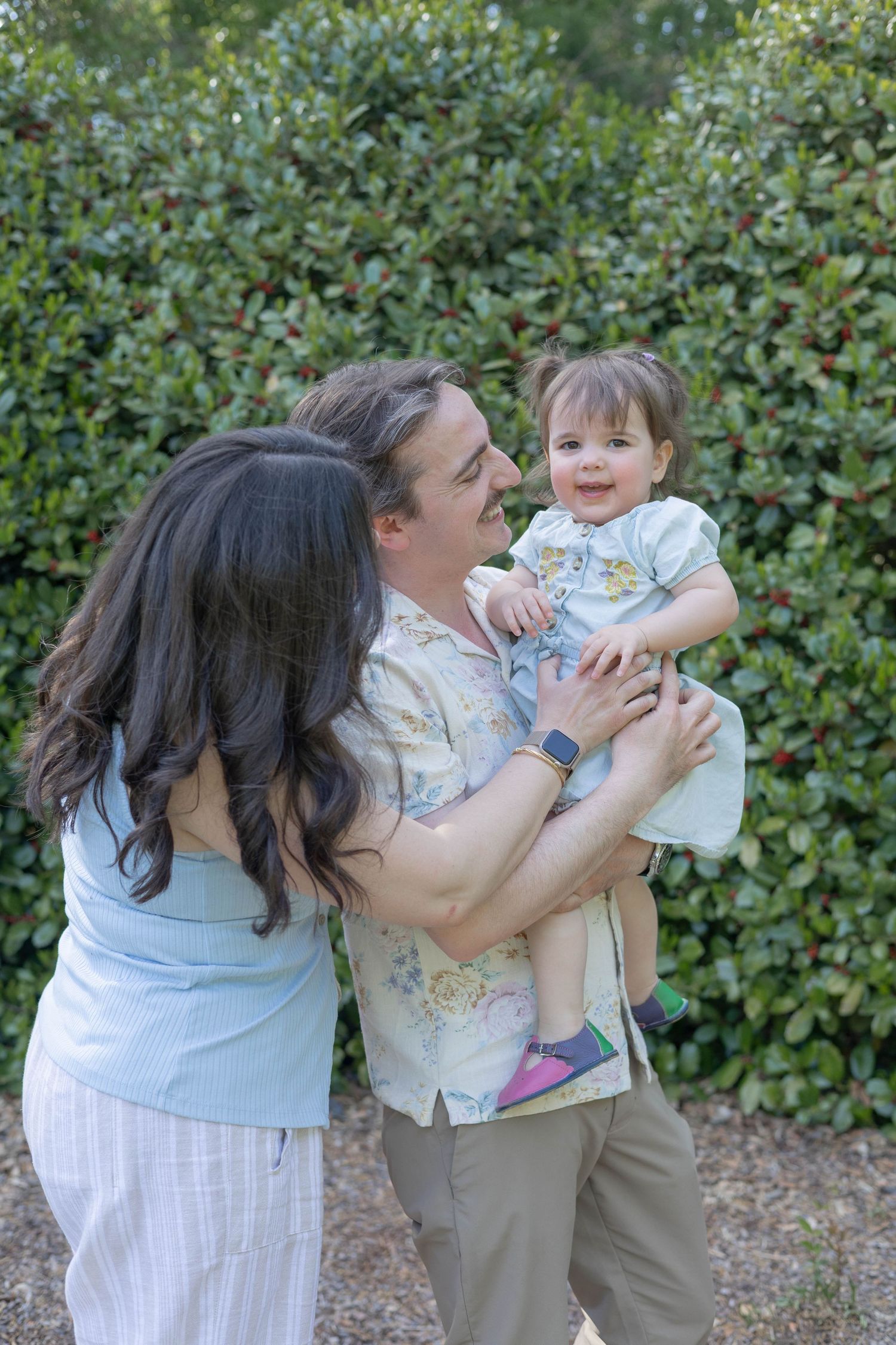 Happy family with parents holding smiling toddler outdoors in front of lush green hedges in Pinehurst NC.