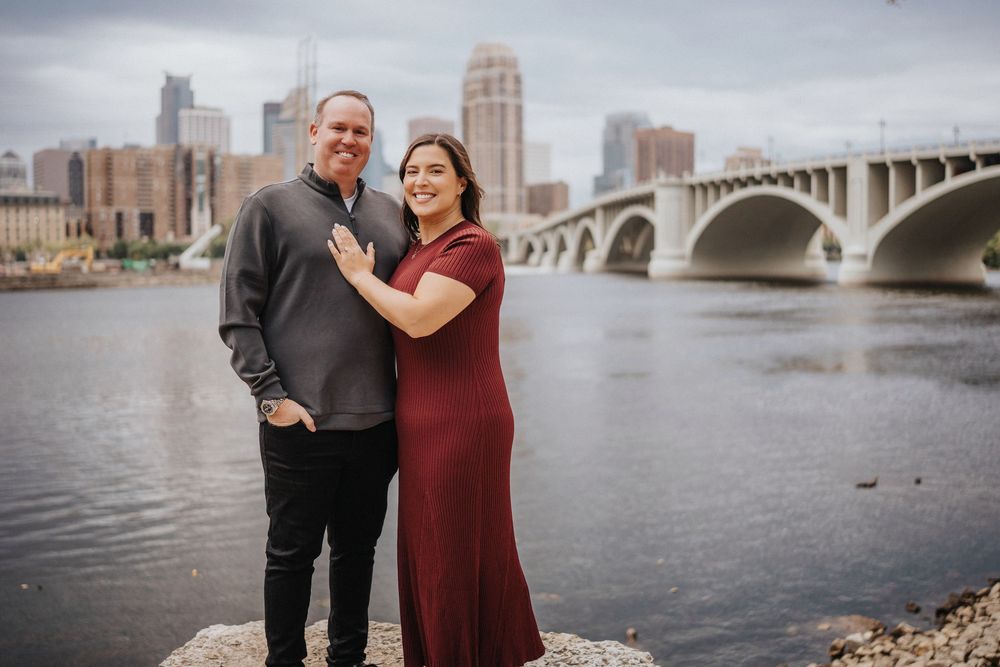 Couple poses together by river with stone bridge and city skyline in background during autumn engagement photoshoot.