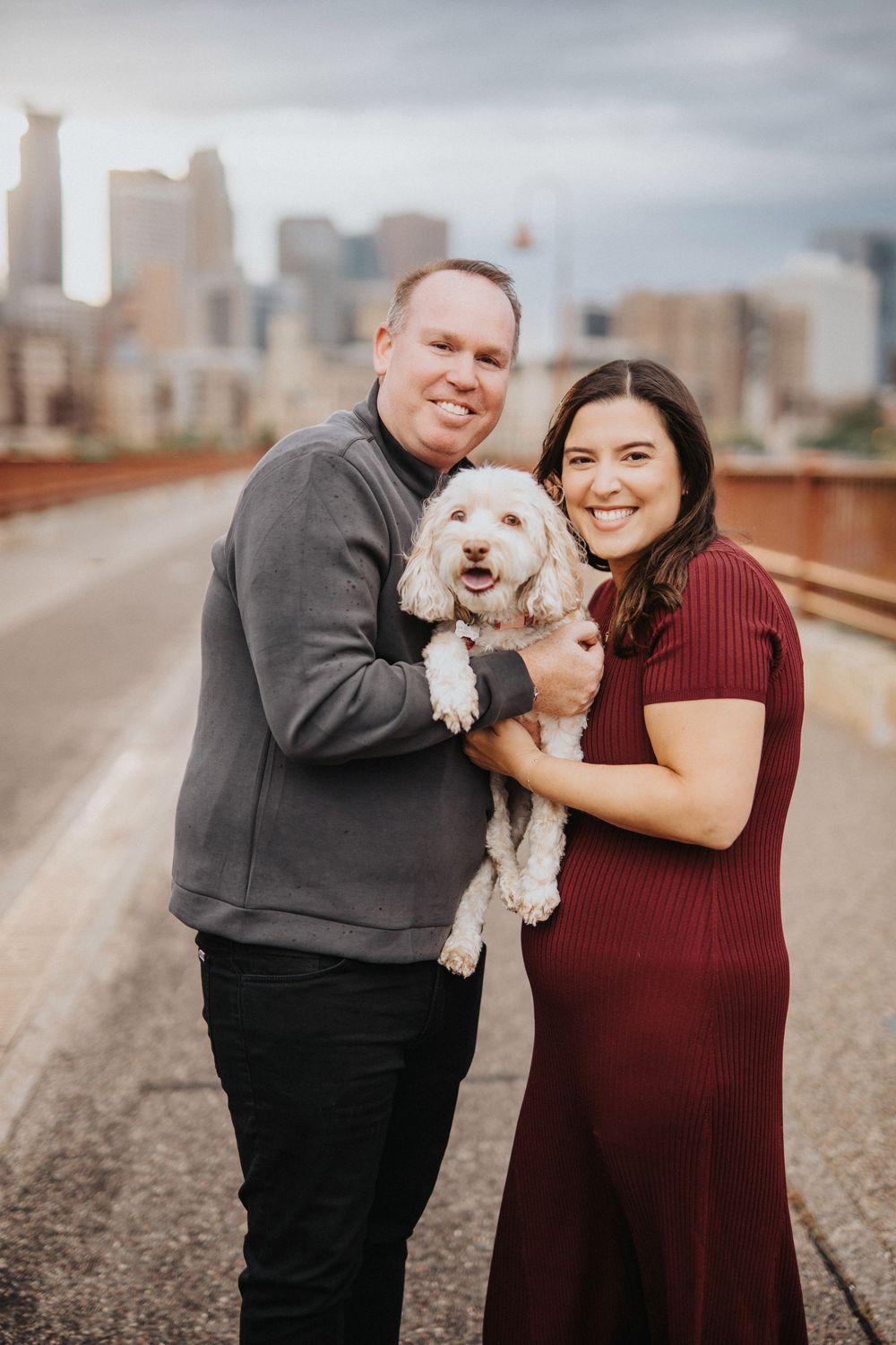 A couple poses with their small white dog on a bridge with a city skyline in the background.