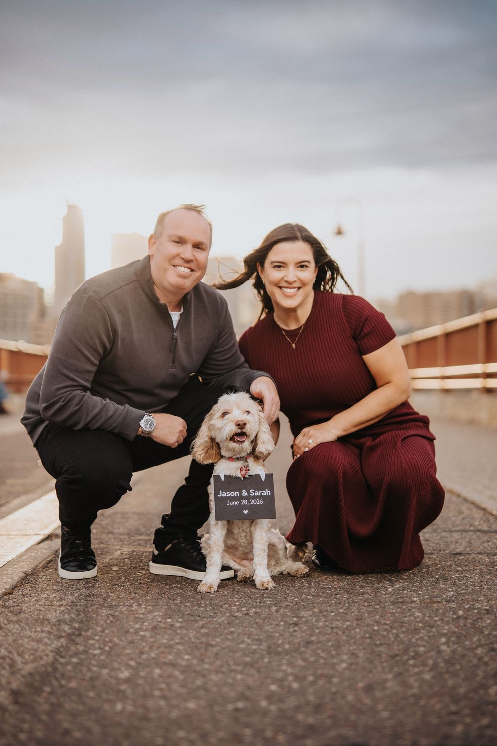 A couple poses playfully with their small white dog on a rooftop with a city skyline in the background.